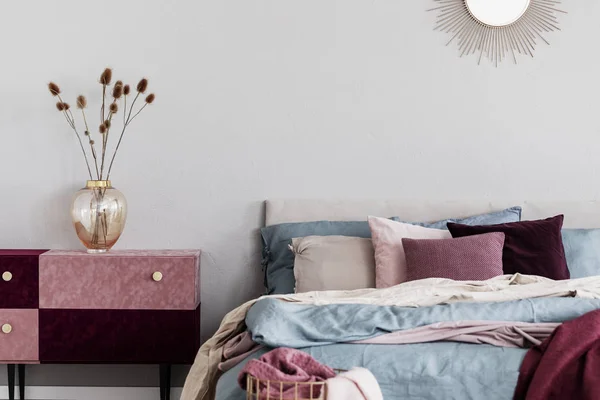 Flores em vaso de vidro elegante em veludo coberto pastel rosa e mesa de cabeceira borgonha ao lado da cama acolhedora com roupa de cama azul e bege — Fotografia de Stock