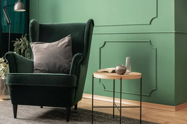 Close-up of a green velvet armchair with a gray cushion standing next to a wooden coffee table in a cozy living room interior with molding on the wall. Real photo — Stock Photo, Image