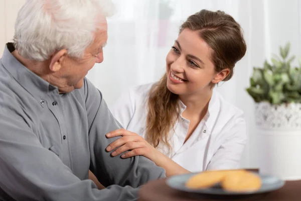 Улыбающаяся молодая медсестра сидит за столом со старшим пациентом — стоковое фото