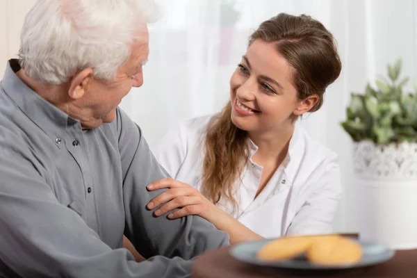 Smiling young nurse sitting at table with senior patient — Stock Photo, Image