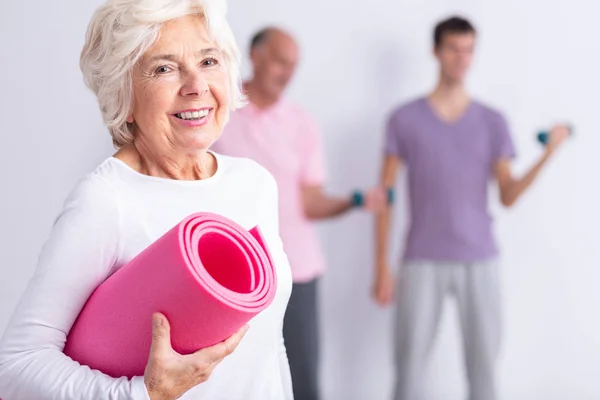 Glad senior kvinna med rosa joga matta, män som tränar i bakgrunden — Stockfoto