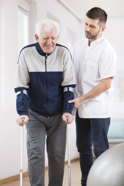 Elderly man walking on crutches and a helpful male nurse supporting him — Stock Photo, Image