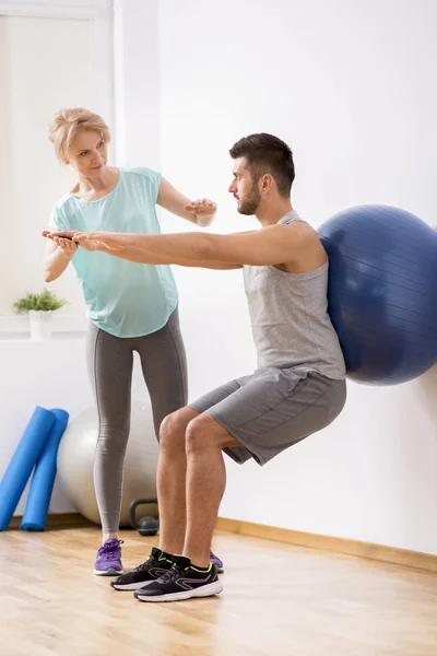 Hombre joven con lesión en la espalda haciendo ejercicio con pelota gimnástica azul durante la cita con la fisioterapeuta femenina — Foto de Stock