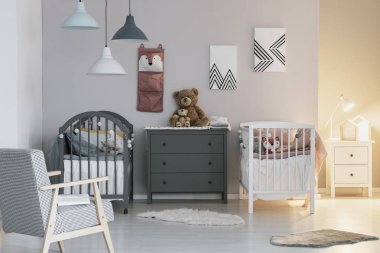 İkiz için moda yatak odasında iki beşik arasında gri ahşap komodin üzerinde kahverengi oyuncak ayı