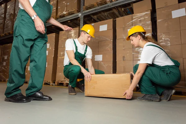 Two young warehouse workers in dark green uniforms and yellow helmets lifting heavy package — Stock Photo, Image