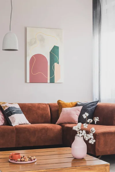 Цветы в вазе на деревянном кофейном столике в модном интерьере гостиной с коричневым угловым диваном с подушками и абстрактной росписью на стене — стоковое фото