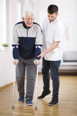 Yaşlı adam koltuk değneği ve yardımcı bir erkek hemşire onu destekleyen yürüyüş