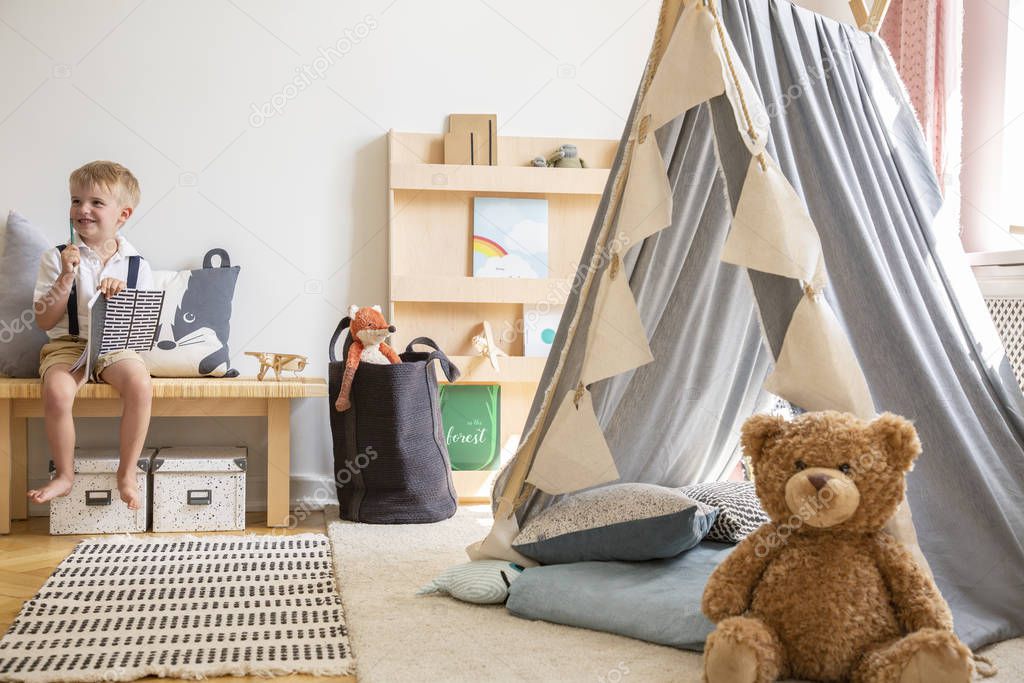 Smiling kid sitting on bench with notebook, real photo of natural playroom interior with scandinavian tent and teddy bear