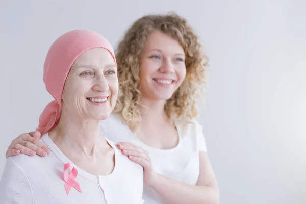 Positieve Senior moeder lijdt aan lupus dragen roze hoofddoek en lint, dochter staande achter haar steun haar — Stockfoto