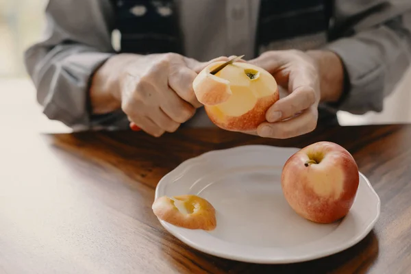 Senior Kaukasische man peeling een appel met een mes zittend aan de houten tafel — Stockfoto