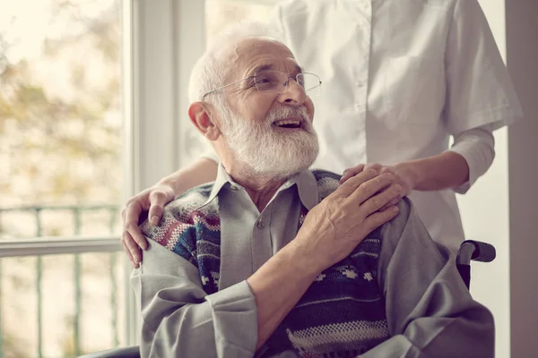 Senior man zittend op de rolstoel, lachen en houden van de hand van zijn verpleegkundige — Stockfoto