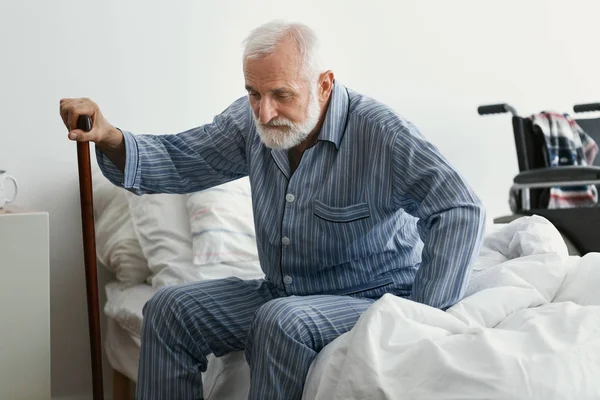 Грустный пожилой человек с болезнью Альцгеймера держит трость и сидит на кровати в доме престарелых — стоковое фото