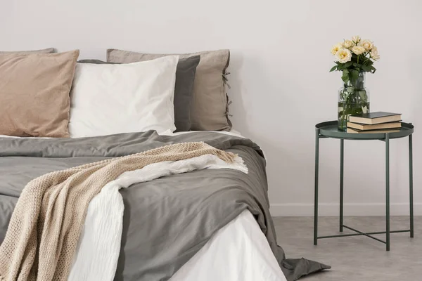 White roses in glass vase and books on stylish nightstand next to big bed with linen pillows and cozy blanket — Stock Photo, Image