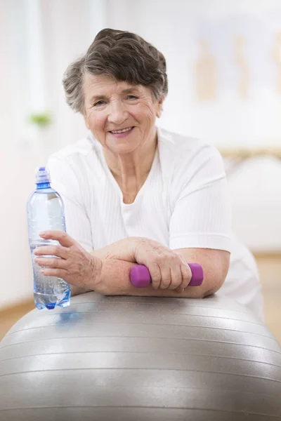 Старшая женщина с бутылкой воды и гантели опираясь на серый гимнастический мяч — стоковое фото