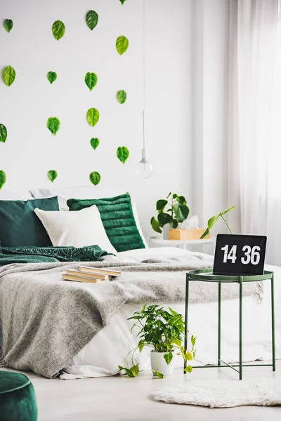Laptop su tavolino verde metallo davanti a letto accogliente con cuscino smeraldo e coperta grigia, foglie sulla parete bianca — Foto Stock