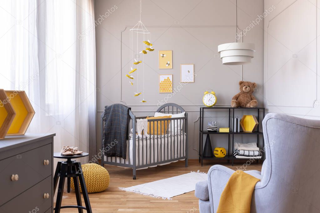 Dark grey blanket on wooden crib in yellow and grey baby bedroom with armchair and industrial metal shelf with toys