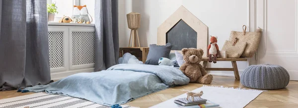 Panoramisch uitzicht op de kinder slaapkamer en speelkamer met een klein bed met blauw beddengoed, teddybeer op de vloer en boeken op het tapijt — Stockfoto