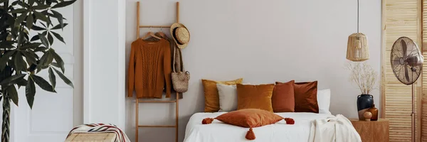 Almohada en una cama junto a una escalera con jerséis colgantes en el interior de un dormitorio natural — Foto de Stock