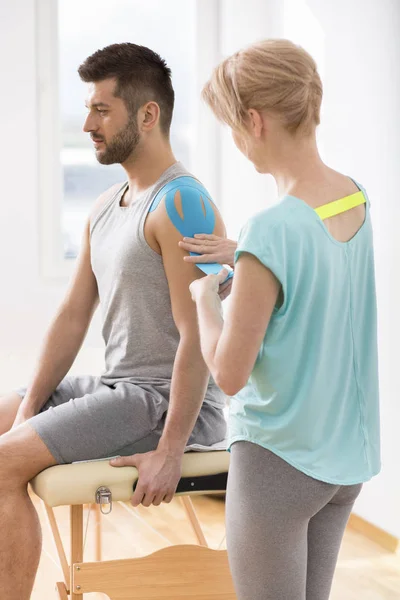 Fysiotherapeut die geselecteerde fragmenten van het lichaam van de jonge man bedekt met speciale structuur patches tijdens kinesiotaping therapie — Stockfoto