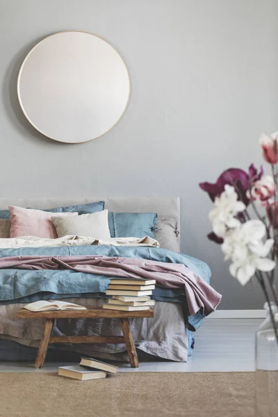 Round mirror in wooden frame on grey wall of elegant bedroom interior with comfortable bed with pastel, blue and pink bedding