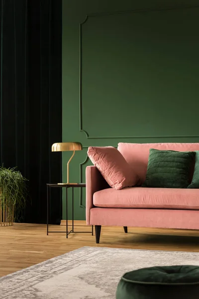 Stylish golden lamp on industrial table next to pink couch with emerald pillows in dark green living room interior, copy space on empty wall — стоковое фото