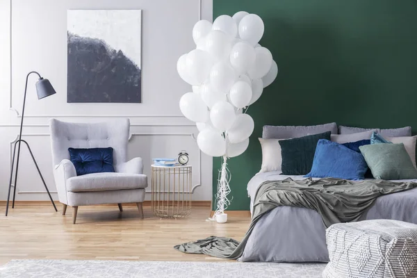 Ballons blancs dans un élégant intérieur de chambre à coucher avec fauteuil confortable gris et lit chaud, vraie photo avec espace de copie sur le mur gris vide — Photo