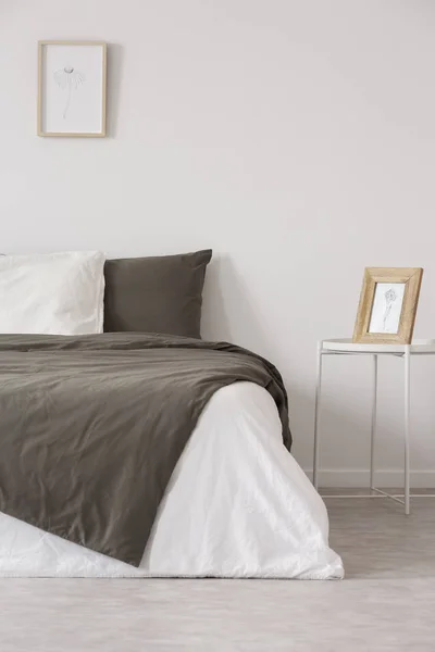 Gráfico en marco de madera sobre mesita de noche de metal blanco junto a cómoda cama con almohadas negras y grises y edredón —  Fotos de Stock