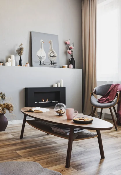 Kekse und rosa Teetasse auf Holztisch im grauen Wohnzimmerinterieur mit Öko-Kamin und schickem Stuhl — Stockfoto