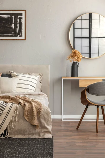 Elegant round mirror in wooden frame above fancy console table with flowers in vase in trendy bedroom interior with beige vase