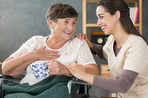 Femme âgée positive en fauteuil roulant tenant une tasse de thé et souriant à un beau bénévole à la maison de soins infirmiers — Photo