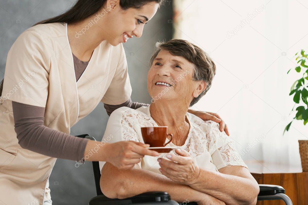 Pretty smiling nurse is giving herbal tea to positive elderly lady in a wheelchair at nursing home