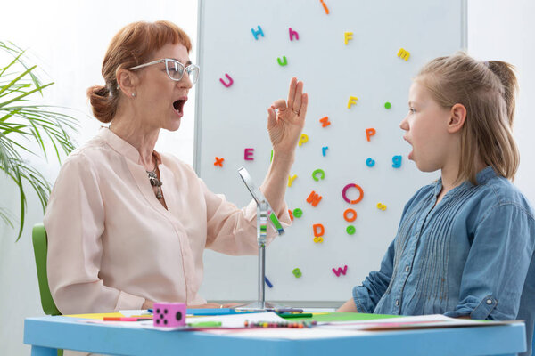 Милая блондинка, открывающая рот во время логопедии со старшим профессиональным терапевтом, копирует пространство на стене буквами
