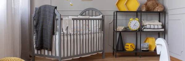 Panoramic view of cute yellow and grey baby bedroom with grey crib and black shelf with book, toys, blankets and clock — Stock Photo, Image