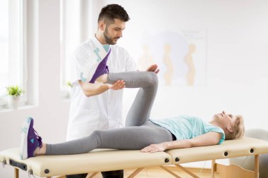 Genç yakışıklı doktor ile oturum sırasında fizyoterapi masasında yatan diz yaralanması ile orta yaş kadın