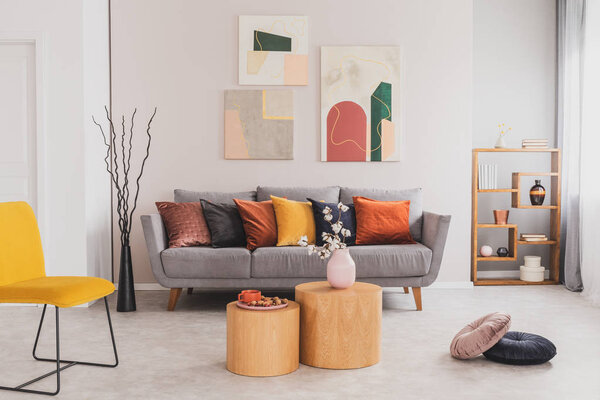 Yellow, orange, black and brown pillows on comfortable grey scandinavian sofa in bright living room interior with abstract paintings on the wall