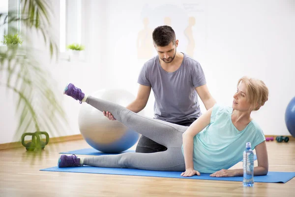 Middle age woman exercising on blue mat during physiotherapy with young male doctor — Stock Photo, Image
