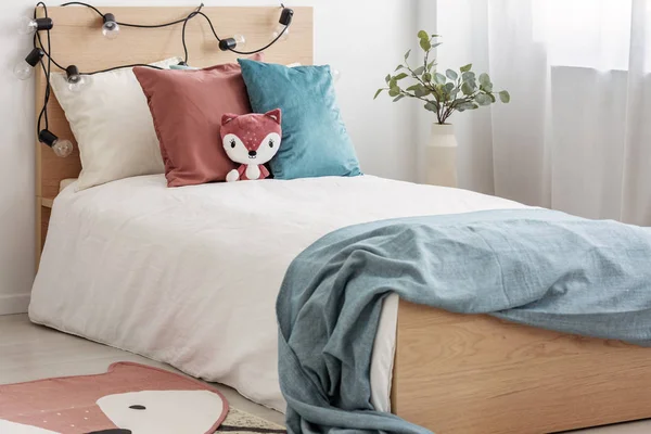 Бархатная пастельно-розовая и бирюзовая подушки и игрушечная лиса на односпальной деревянной кровати с белым постельным бельем и одеялом — стоковое фото