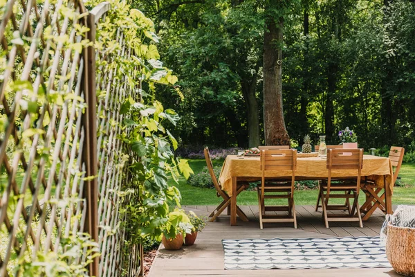 Mesa de comedor cubierta con mantel naranja de pie en terraza de madera en jardín verde — Foto de Stock