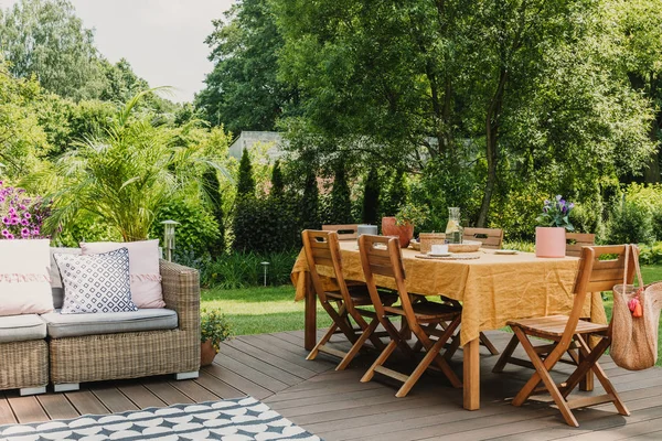 Τραπέζι φαγητού καλυμμένο με πορτοκαλί τραπεζομάντηλο που στέκεται σε ξύλινη βεράντα στον καταπράσινο κήπο που προετοιμάζεται για πάρτι κήπου — Φωτογραφία Αρχείου