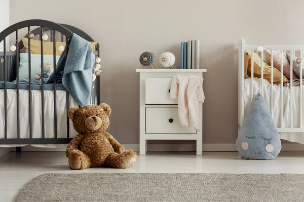 Urso de pelúcia marrom no chão de madeira do interior do quarto de bebê escandinavo com mesa de cabeceira branca e berços — Fotografia de Stock