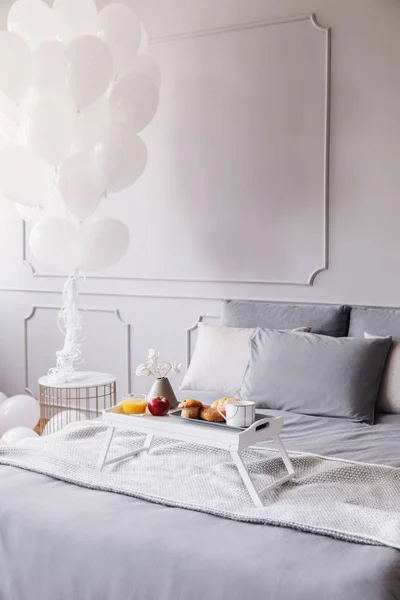 Petit déjeuner d'anniversaire sur trey en bois blanc sur grand lit avec draps gris et couverture, bouquet de ballons blancs au-dessus de la table de nuit, espace de copie sur le mur vide — Photo