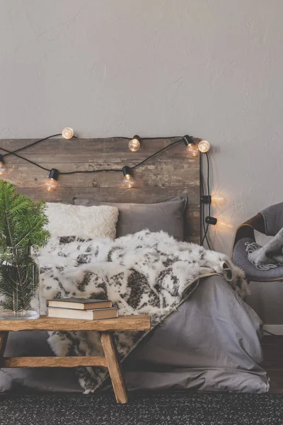 Christmas tree in glass vase next to books on wooden bench in the foot of a warm king size bed with wooden headboard — Stock Photo, Image