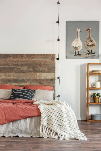 Strickdecke auf einem Bett mit hölzernem Bettkopf vor weißer Wand mit einem Ölgemälde von Enten. echtes Foto des Schlafzimmers — Stockfoto