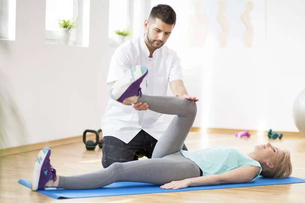 Middle age woman exercising on blue mat during physiotherapy with young male doctor — Stock Photo, Image