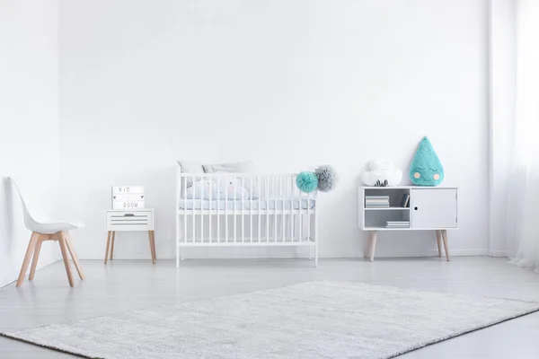 Cojín azul en el gabinete junto a la cuna en el interior del dormitorio del bebé blanco con silla y alfombra. Foto real —  Fotos de Stock