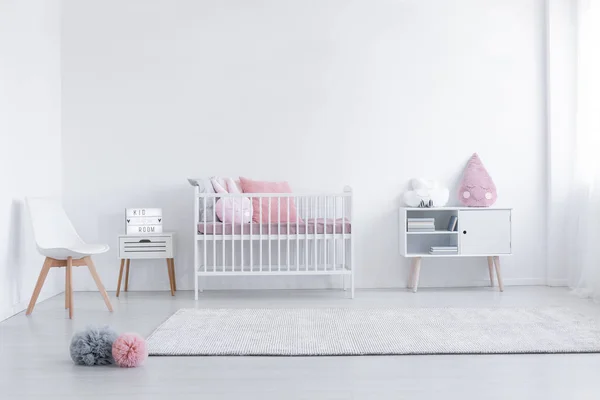Roze kussen op kast en witte stoel in eenvoudige kinderkamer interieur met tapijt en wieg. Echte foto — Stockfoto