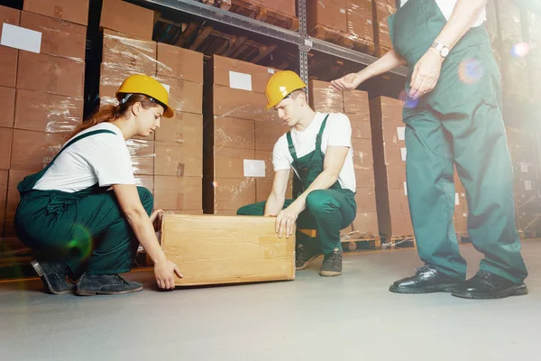 Zwei junge Lagerarbeiter in dunkelgrünen Uniformen und gelben Helmen heben schweres Paket — Stockfoto