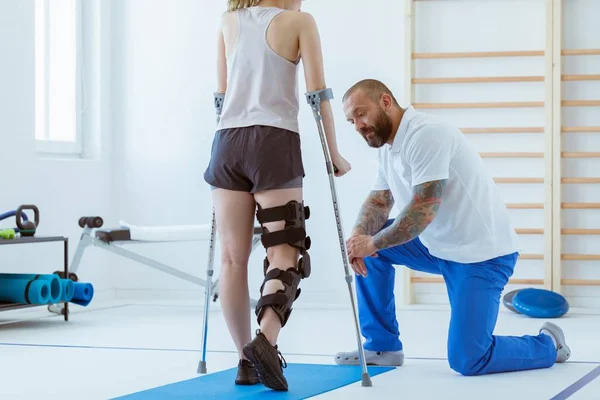 Koltuk değneğinde ortozu olan bir kız hastanedeki bir spor salonunda mavi bir paspas üzerinde egzersiz yapıyor. — Stok fotoğraf