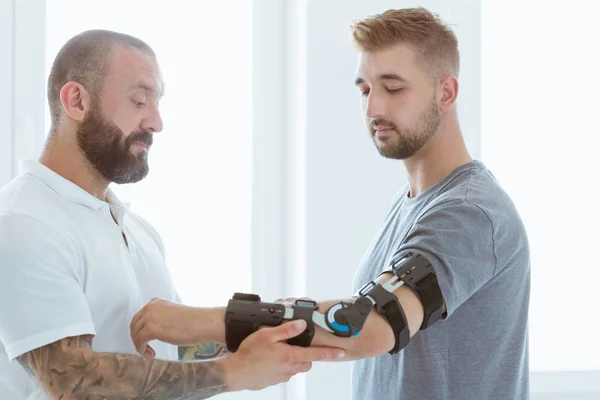 Tattooed physiotherapist who puts an orthosis on the hand of a young patient after an accident — Stock Photo, Image