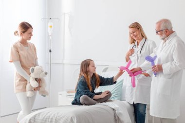 İki doktor hastane yatağında balon hayvanlarla küçük kızı neşelendiriyorlar.